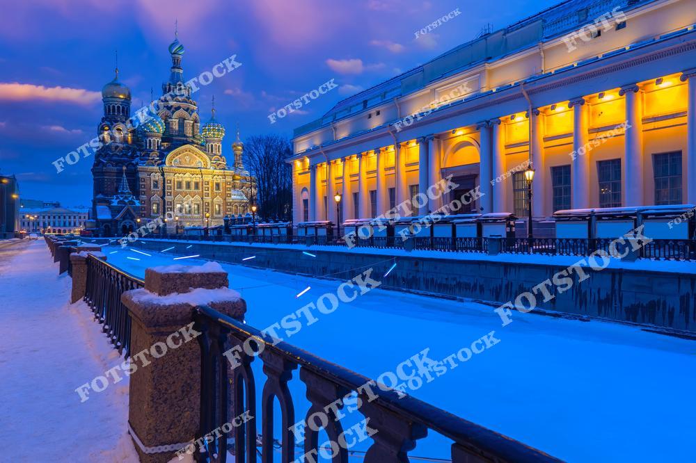На фотографии изображён храм Спаса-на-Крови, один из самых узнаваемых символов Санкт-Петербурга. Он возвышается над заснеженным каналом Грибоедова, вода в котором покрыта льдом. Справа — колоннада Русского музея, красиво освещённая в вечернее время. Снег покрывает тротуары и ограждения, создавая атмосферу спокойствия и зимней тишины. Вечерний свет подчёркивает архитектурные детали и купола храма.